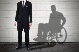 Man in a suit standing against a brick wall, with his shadow revealing the silhouette of a person in a wheelchair, symbolizing hidden disabilities or overcoming challenges.