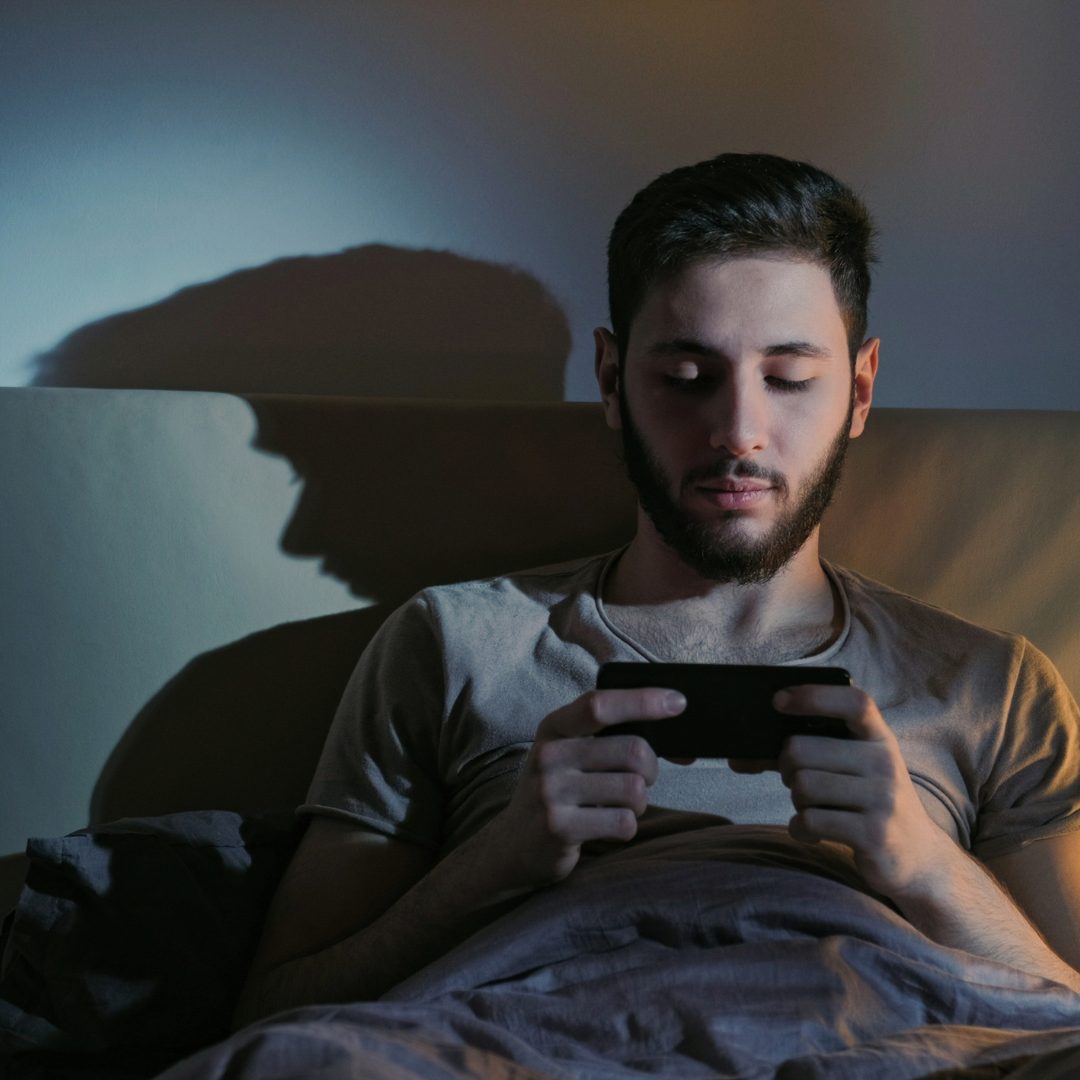A young man with dark hair and a beard sitting in bed, illuminated by the glow of a smartphone he is holding with both hands. The background shows a plain wall with a shadow cast by the phone's screen. The man appears focused on his device in a dimly lit room.