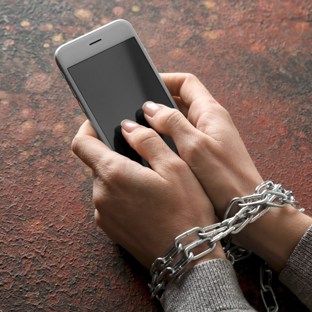 A person holding a smartphone with both hands. Their wrists are chained together with a thick metal chain. The background surface is rough and reddish-brown in color.