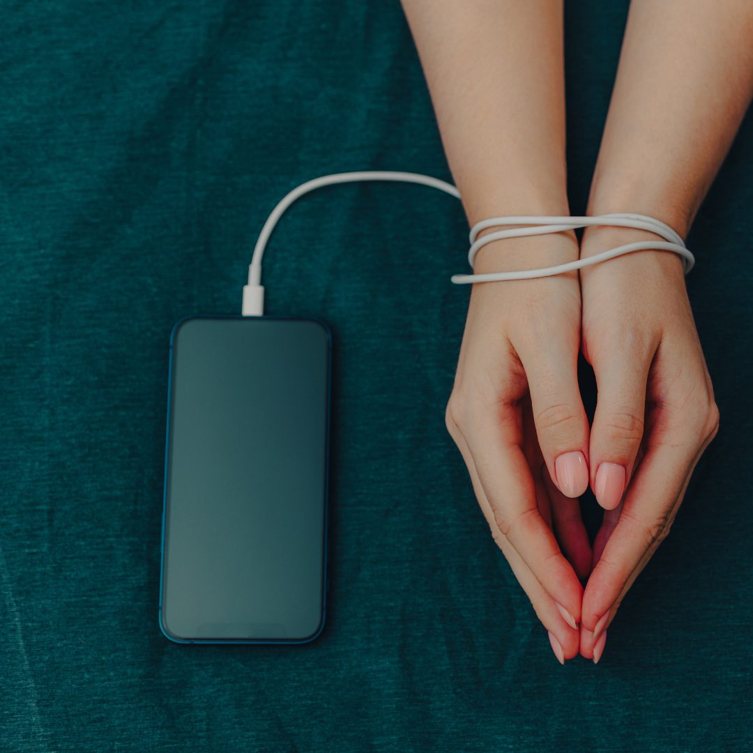 A pair of hands bound together with a white charging cable, connected to a smartphone placed on a teal fabric surface. The person's nails are neatly trimmed and painted with a light pink polish.
