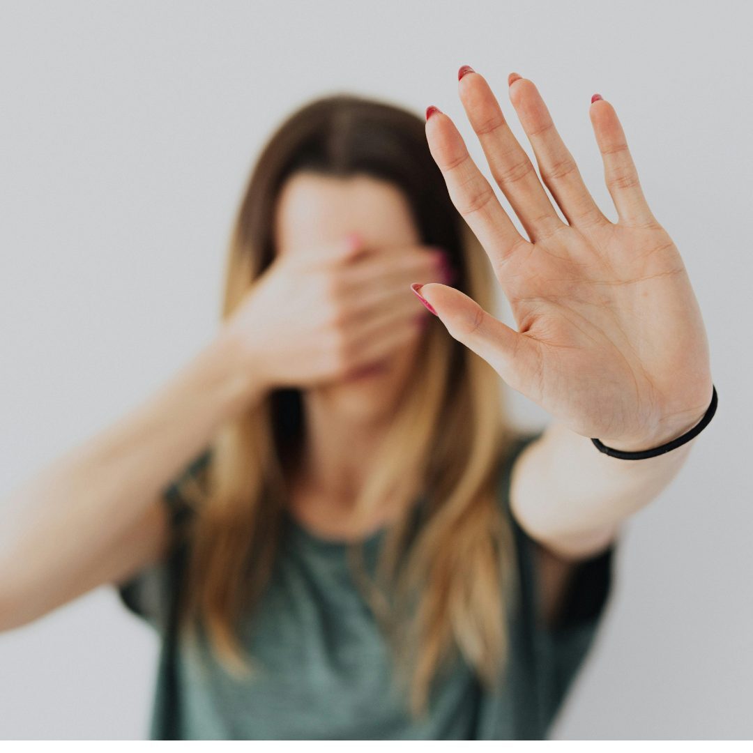 A woman with long, light brown hair is holding up her hand with her palm facing the camera, as if signaling "stop." She is slightly blurred in the background, with her other hand covering her face. The background is plain and light-colored.