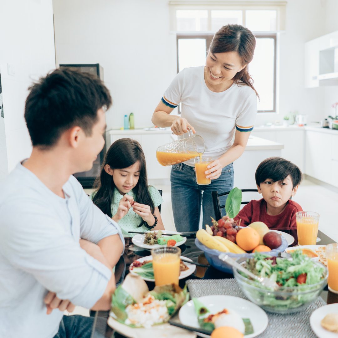 A family gathered around a dining table, enjoying a meal together. A woman is pouring orange juice into a glass, smiling warmly. Two children, a girl and a boy, are seated at the table, with the girl looking at her food and the boy looking off to the side. The table is filled with various foods including fruits, salads, and glasses of orange juice, in a bright kitchen setting.