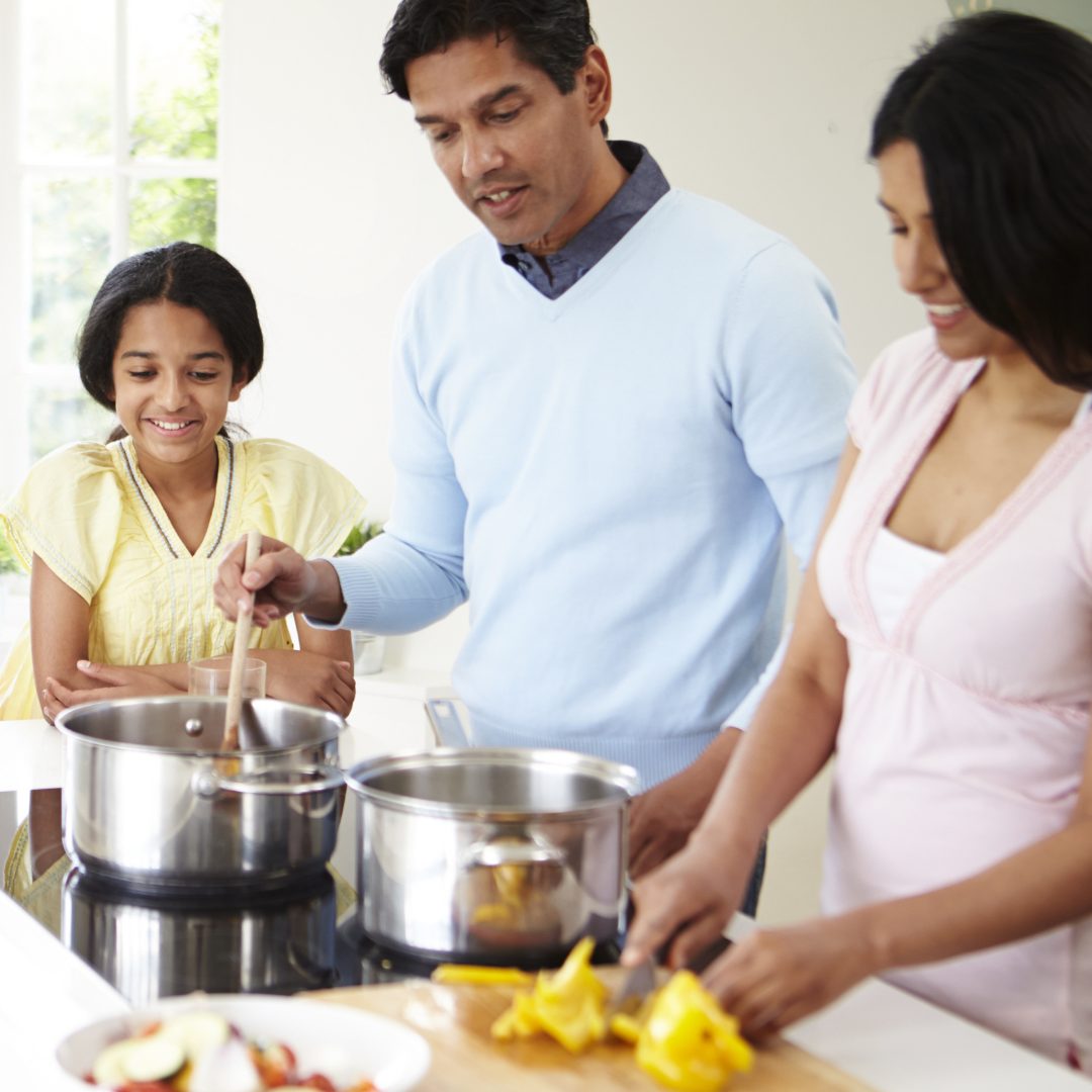 A family gathered in the kitchen, with a man stirring a pot on the stove, a woman chopping yellow bell peppers, and a young girl smiling and watching. The scene is bright with natural light coming through a window.