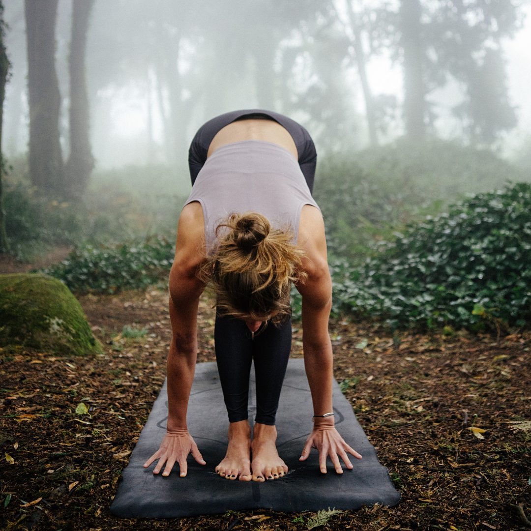 A woman is practicing yoga outdoors on a black mat in a foggy forest. She is in a forward bend pose with her hands on the ground and her head hanging down, her feet together, and her back rounded. She is wearing a gray tank top and black leggings. The background features tall trees and dense greenery shrouded in mist.
