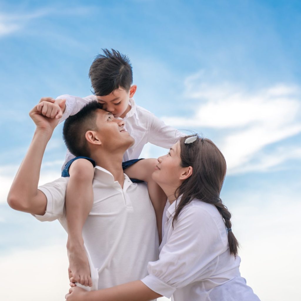 happy family of three outside smiling with a little boy sitting on his dad's head and his mom is hugging his dad.