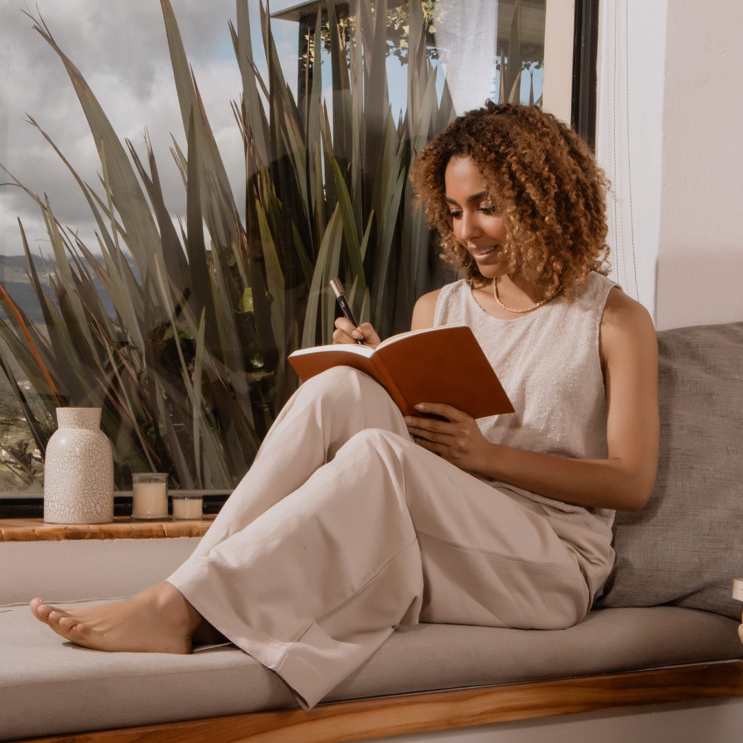 A woman with curly, shoulder-length hair and a warm smile is sitting on a cushioned window seat indoors. She is wearing a sleeveless beige top and light-colored wide-leg pants. She is holding a pen and a notebook, appearing to be writing or sketching. Behind her, there is a large window with greenery outside, and on the windowsill, there are decorative vases and candles.