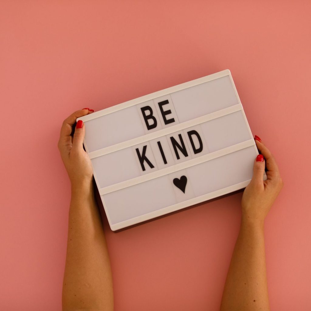 A person with red-painted nails holding a white lightbox against a pink background. The lightbox displays the message "BE KIND" with a small black heart symbol below.