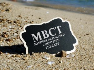Small black sign resting on sandy beach that reads “MBCT Mindfulness-Based Cognitive Therapy,” photographed near the shoreline.