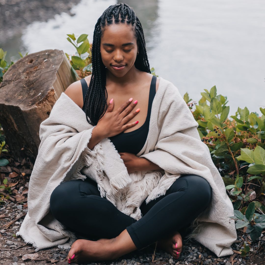 A young woman with braided hair is sitting cross-legged outdoors near a body of water, surrounded by greenery. She has her eyes closed and is gently resting her right hand on her chest, with a peaceful expression on her face. She is wearing a black top, black pants, and a light-colored shawl draped over her shoulders.