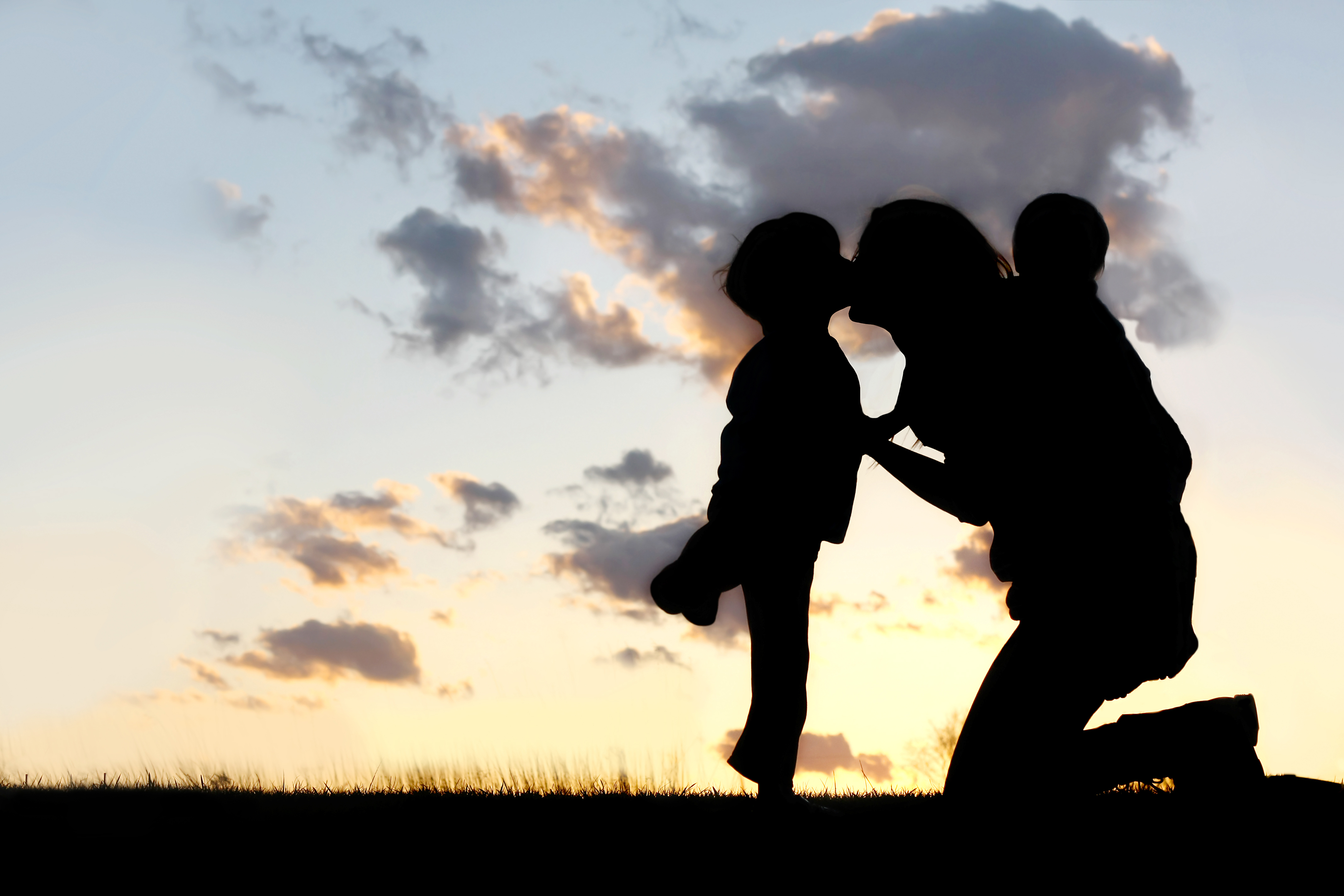 Silhouette of a mother kneeling to kiss her child while holding another child close, set against a soft sunset sky with scattered clouds.