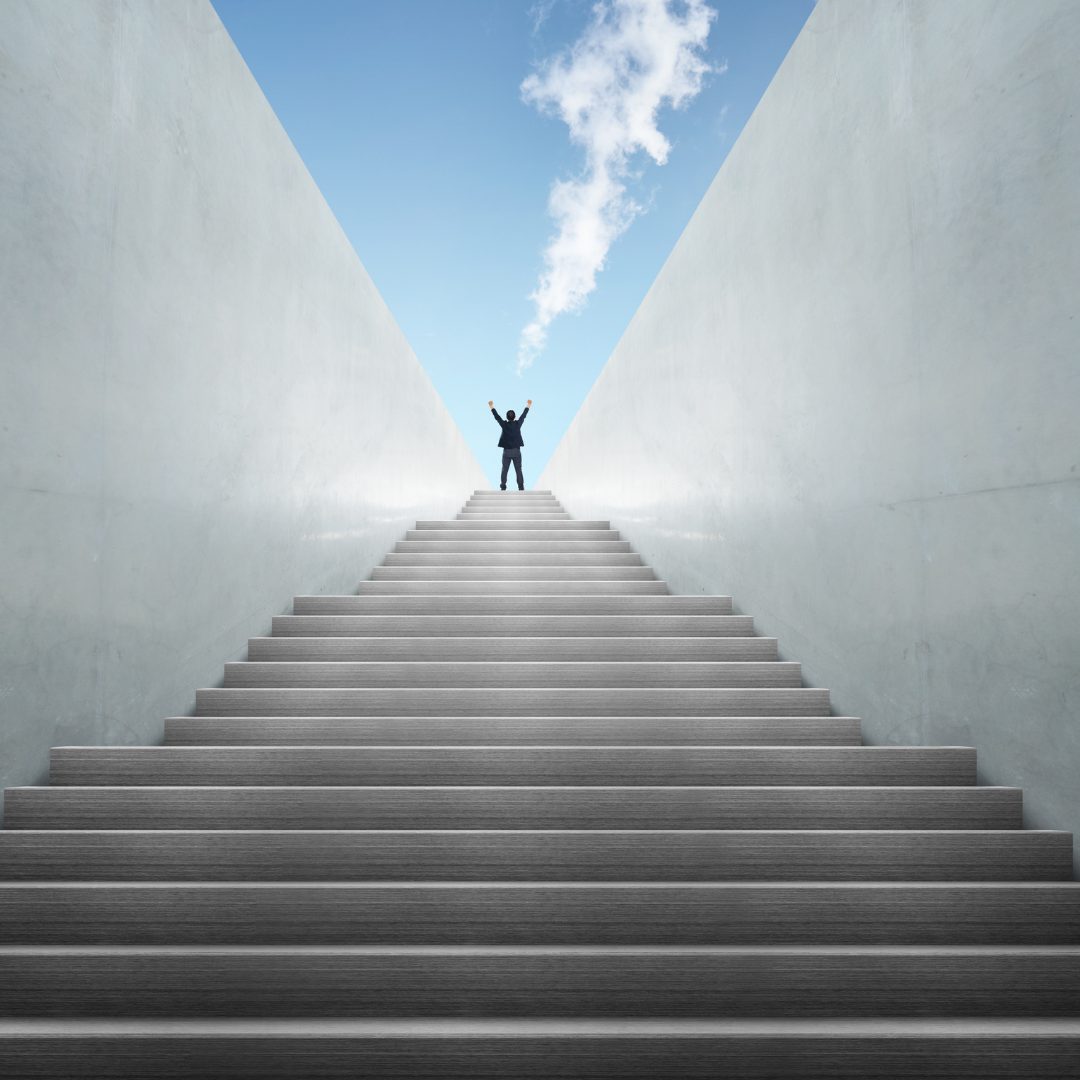 A person dressed in a suit stands at the top of a wide, modern staircase with their arms raised in victory. The staircase is flanked by tall, white walls, and the sky above is blue with some clouds. The person appears to be celebrating or expressing achievement.