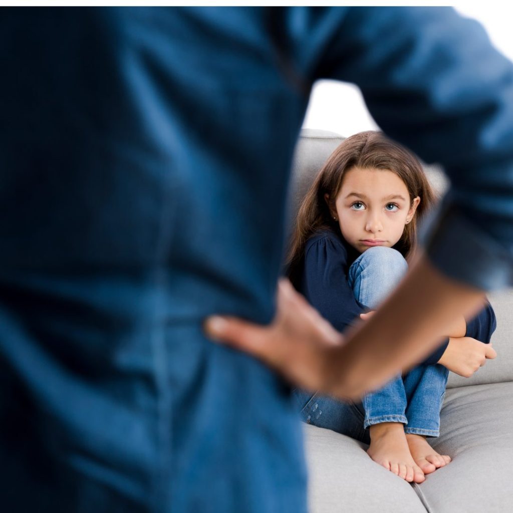 A young girl sitting on a couch, hugging her knees and looking up with a sad or troubled expression. In the foreground, an adult figure with crossed arms is partially visible, creating a sense of distance or confrontation between the two.
