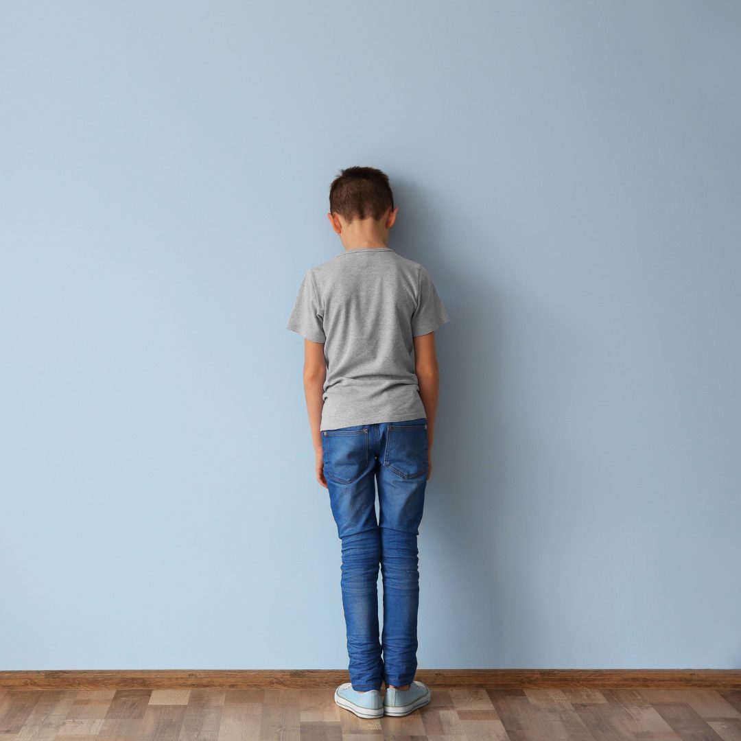 A young boy with short hair, wearing a gray t-shirt and blue jeans, standing with his back against a light blue wall. He appears to be looking down or away, with his arms hanging loosely by his sides. The floor is wooden.
