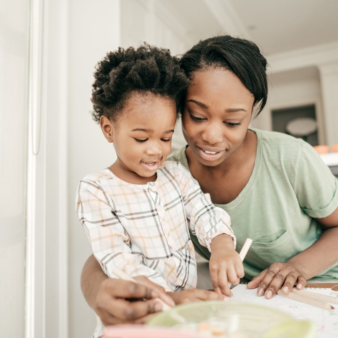 A young woman and a small girl are looking at something together on a table, with the woman smiling and the girl pointing. The woman has short black hair and is wearing a light green shirt, while the girl has curly hair and is wearing a plaid shirt. They appear to be engaged in a shared activity or learning moment.
