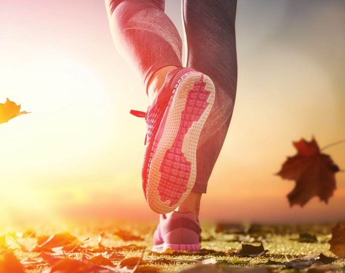 A person wearing athletic shoes is running outdoors on a path covered with fallen autumn leaves. The photo captures the runner's lower legs and feet in motion, with the sun setting in the background, creating a warm, golden glow. Maple leaves are visible falling around the runner, emphasizing the autumn setting.