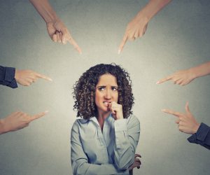 Anxious woman biting her nails while multiple hands around her point accusingly, symbolizing blame, pressure, or overwhelming judgment.