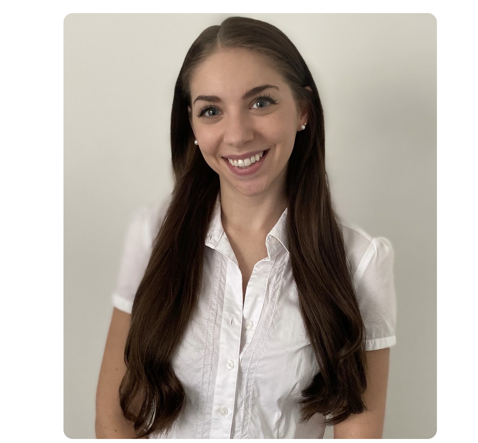 Samantha Rose wearing a long, dark brown hair, styled with a side part, is smiling warmly at the camera. She is wearing a white, short-sleeved, button-up blouse and pearl stud earrings, standing against a plain, light-colored background.