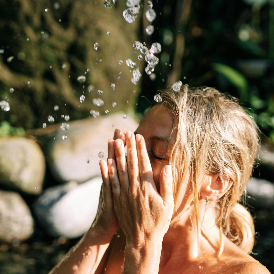 A woman with wet, blonde hair is outdoors, with her eyes closed and hands gently covering her face. Water droplets are falling around her, suggesting she is rinsing or splashing water on herself. The background features blurred greenery and rocks.