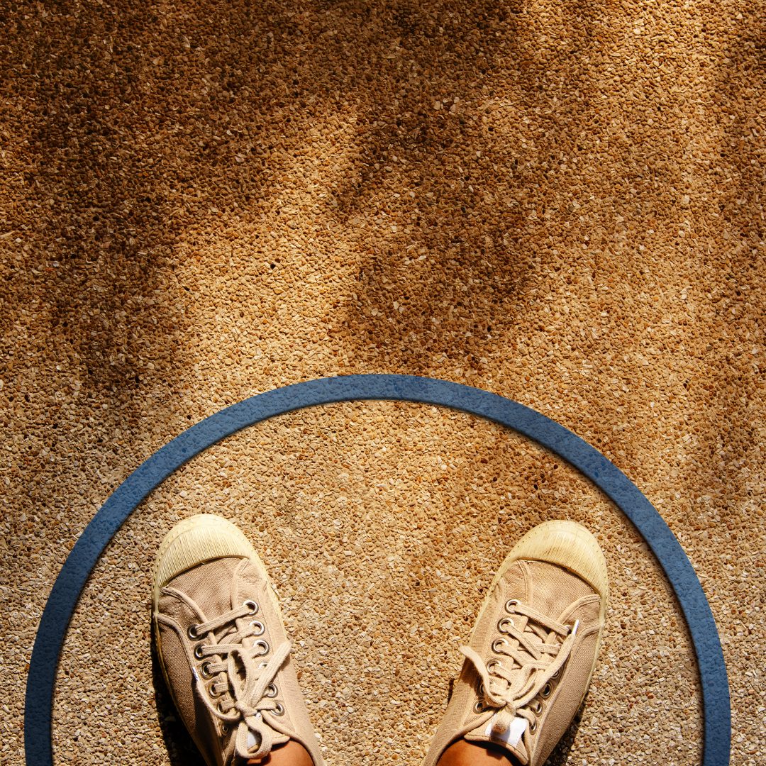 A pair of beige sneakers standing on a sandy or gravelly ground, viewed from above, with a blue circular line partially encircling the shoes.