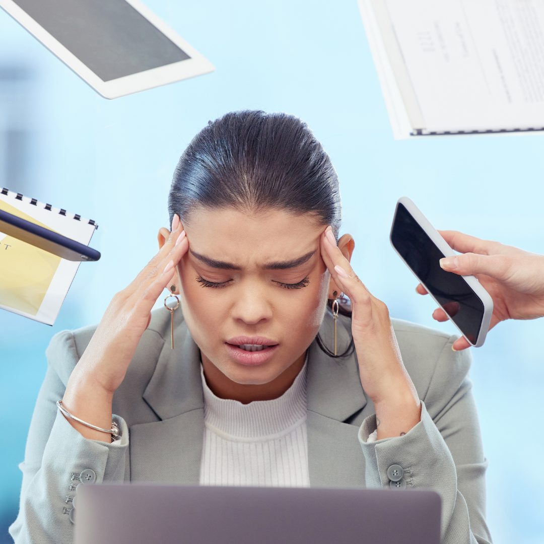 A woman appears stressed and overwhelmed, holding her head with both hands while sitting at a desk with a laptop. Several floating objects around her include a smartphone, notebooks, and documents, suggesting she is dealing with multiple work-related pressures.