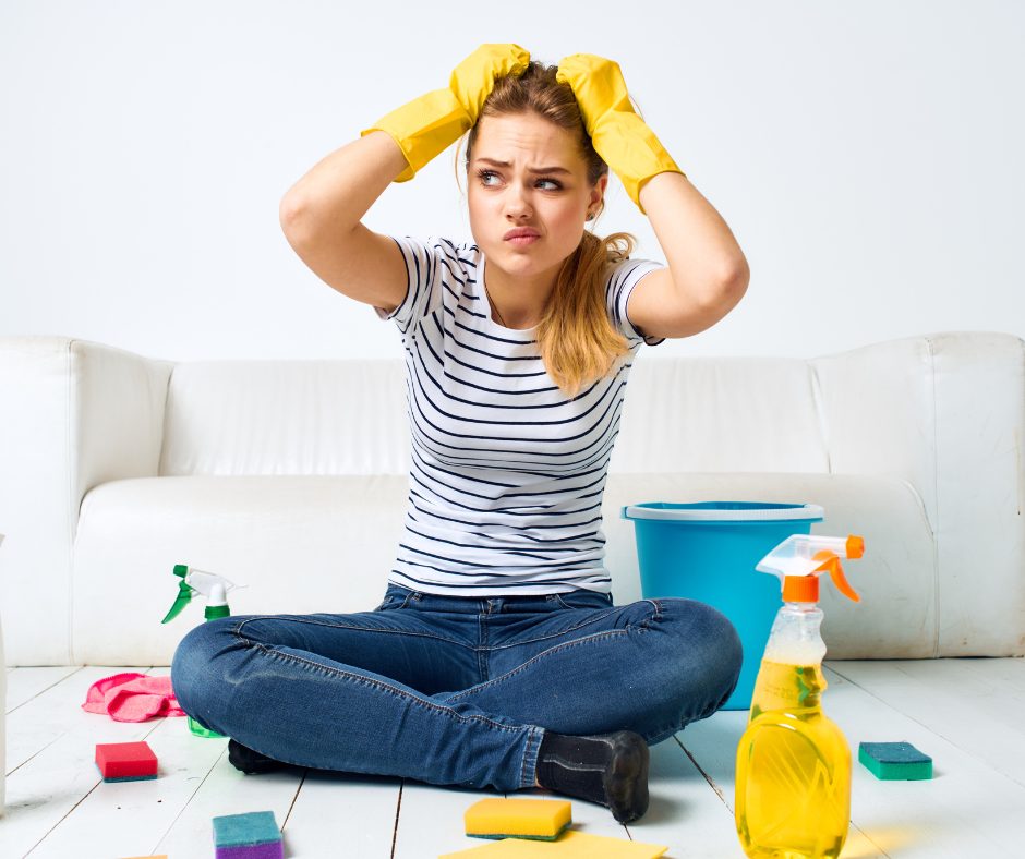 A young woman sitting cross-legged on the floor, wearing a striped t-shirt and blue jeans, with a frustrated or stressed expression as she holds her head and hair with both hands. She is surrounded by cleaning supplies, including spray bottles, sponges, and a bucket, indicating she is in the process of cleaning. The background is plain white, and she is seated in front of a white couch.