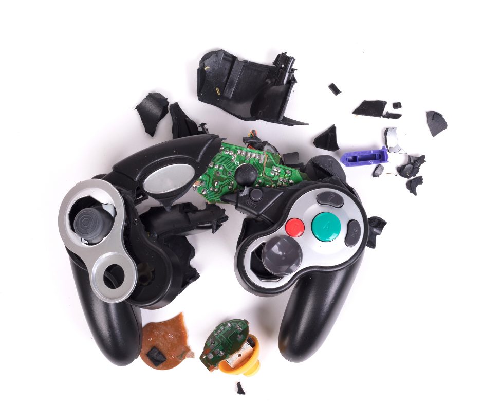 The image shows a pair of disassembled video game controllers with their internal components exposed and broken into pieces. The controllers are black and gray with colorful buttons, and various parts, including circuit boards, plastic fragments, and internal mechanisms, are scattered on a white background.