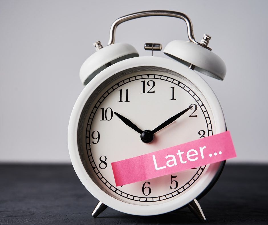 A white analog alarm clock showing the time as 10:10, with a pink sticky note across the bottom that says "Later...". The clock is placed on a dark surface against a plain grey background.