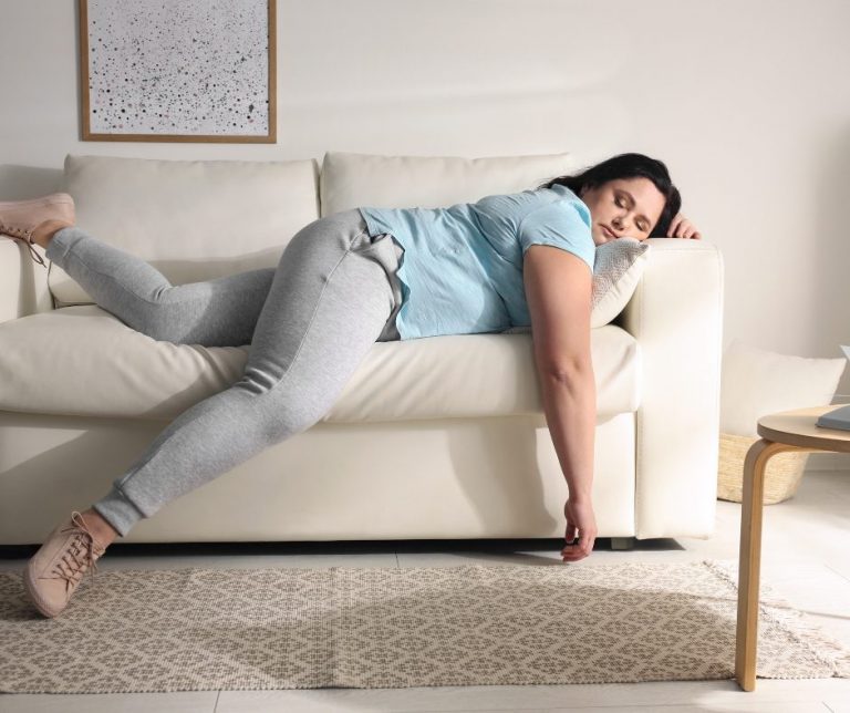 A woman is lying on a white sofa, draped over the armrest with her head resting on a pillow, appearing tired or exhausted. She is wearing a light blue t-shirt, gray sweatpants, and beige sneakers. The sofa is in a modern, minimalistic living room with a framed artwork on the wall, a beige rug on the floor, and a small wooden side table nearby.