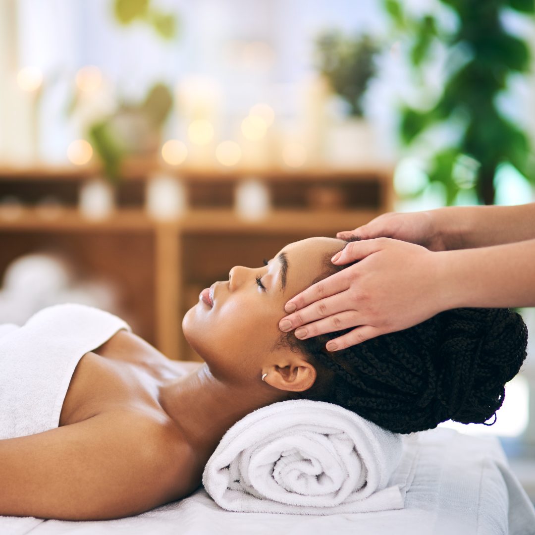 A woman with braided hair is lying on a massage table with her eyes closed, receiving a head massage. She is covered with a white towel, and her head rests on a rolled-up white towel. The background features a spa setting with soft lighting and blurred greenery.