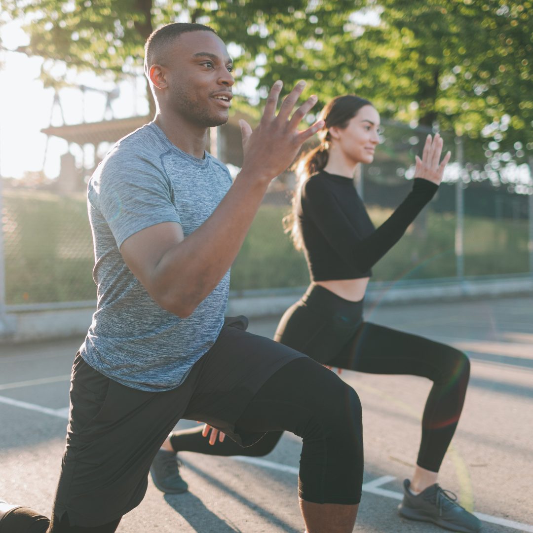 The image shows a man and a woman exercising outdoors on a tennis court, performing lunges with their hands raised in front of them. The man is wearing a gray t-shirt and black shorts, while the woman is dressed in a black long-sleeve top and black leggings. The background features trees and sunlight filtering through the leaves.