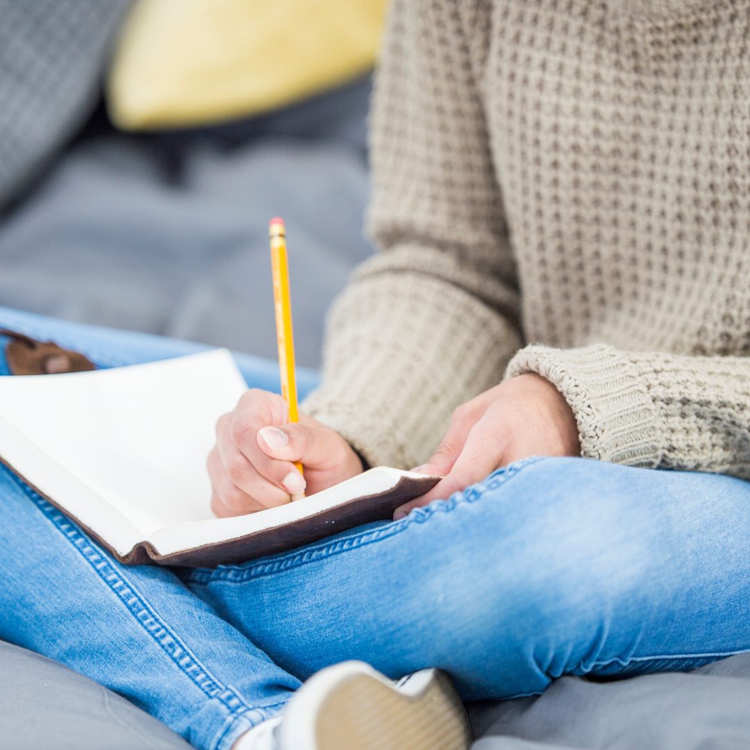 The image shows a person sitting cross-legged on a bed or couch, wearing a beige knit sweater and blue jeans, holding a yellow pencil in one hand and writing or drawing in a blank notebook resting on their lap.