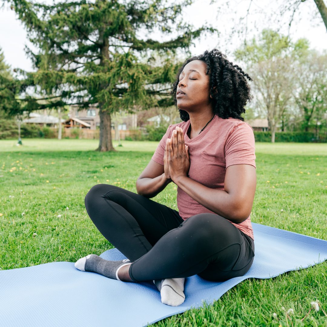 A woman sitting cross-legged on a blue yoga mat outdoors in a park, with her hands pressed together in a prayer position at her chest. She has her eyes closed and a calm expression, practicing meditation or mindfulness. The background features green grass, trees, and a partly cloudy sky.