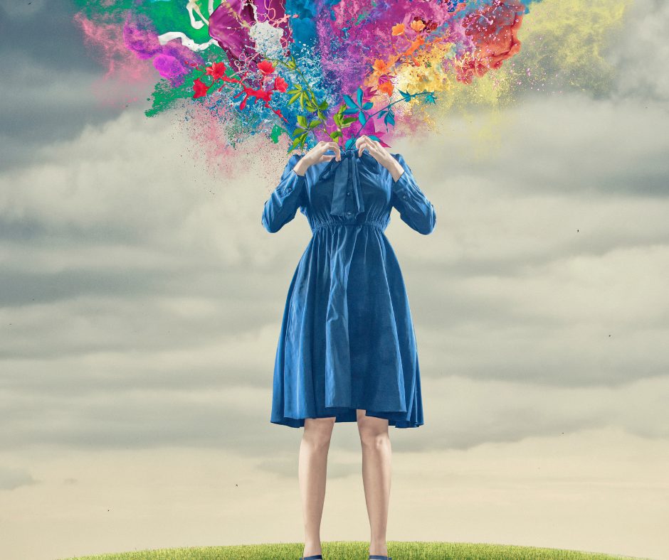 A person standing outdoors on a grassy hill under a cloudy sky, wearing a blue dress. Instead of a head, there is a colorful explosion of paint splashes, flowers, leaves, and abstract shapes emanating upward, creating a vibrant and imaginative visual effect.