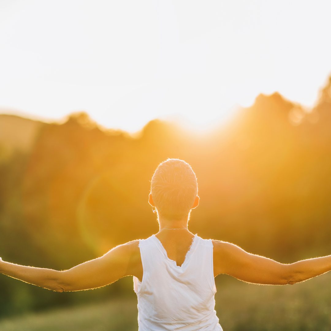 A person with short hair, wearing a sleeveless white top, standing outdoors with arms outstretched, facing the bright sunlight at sunset or sunrise. The background features blurred trees and a glowing sky.