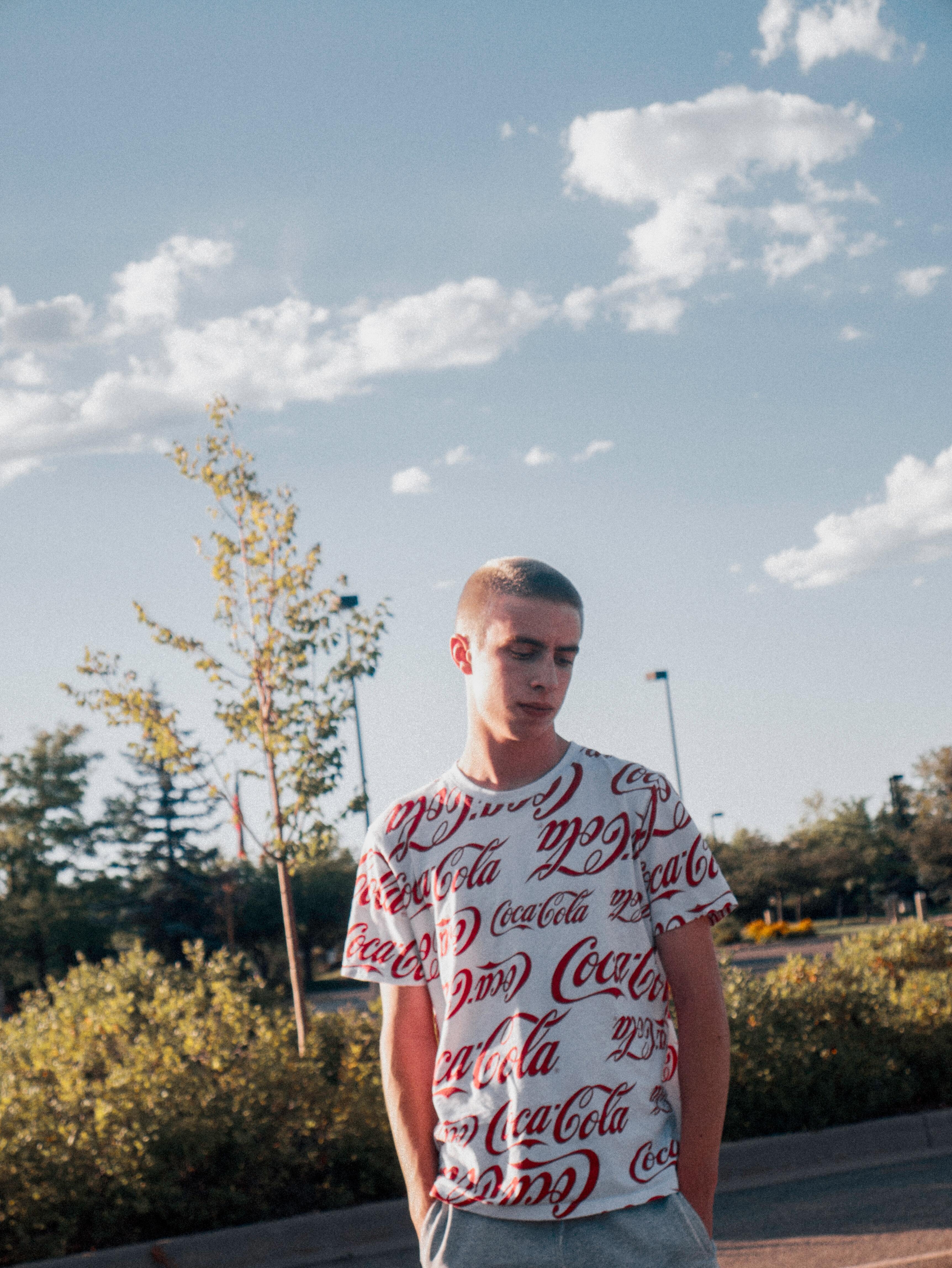 Teenage boy standing outside under a bright sky, wearing a white T-shirt with red Coca-Cola logos, looking down with a thoughtful or introspective expression.