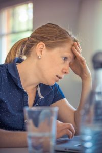 Woman sitting at a desk with her hand on her forehead, staring at her laptop in frustration or worry, with a glass of water in the foreground.