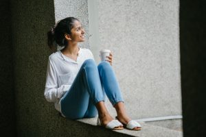 Young woman sitting by a window ledge with her knees up, smiling as she looks outside while holding a cup of coffee, appearing peaceful and content.