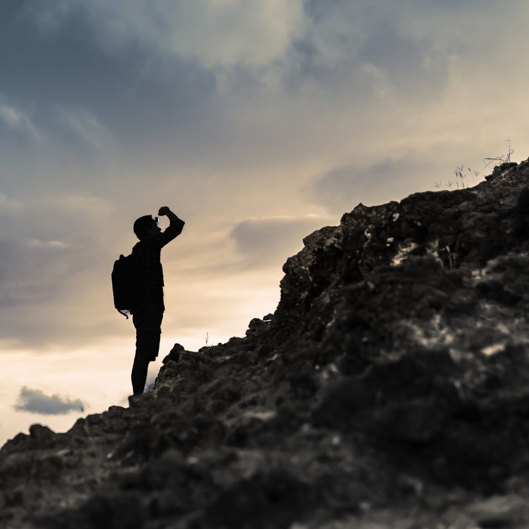 A silhouette of a person with a backpack standing on a rocky slope, drinking from a water bottle, with a cloudy sky in the background during sunset or sunrise.