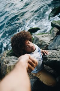Person clinging to coastal rocks near the water, reaching up as another hand pulls them to safety, with a look of urgency and trust on their face.
