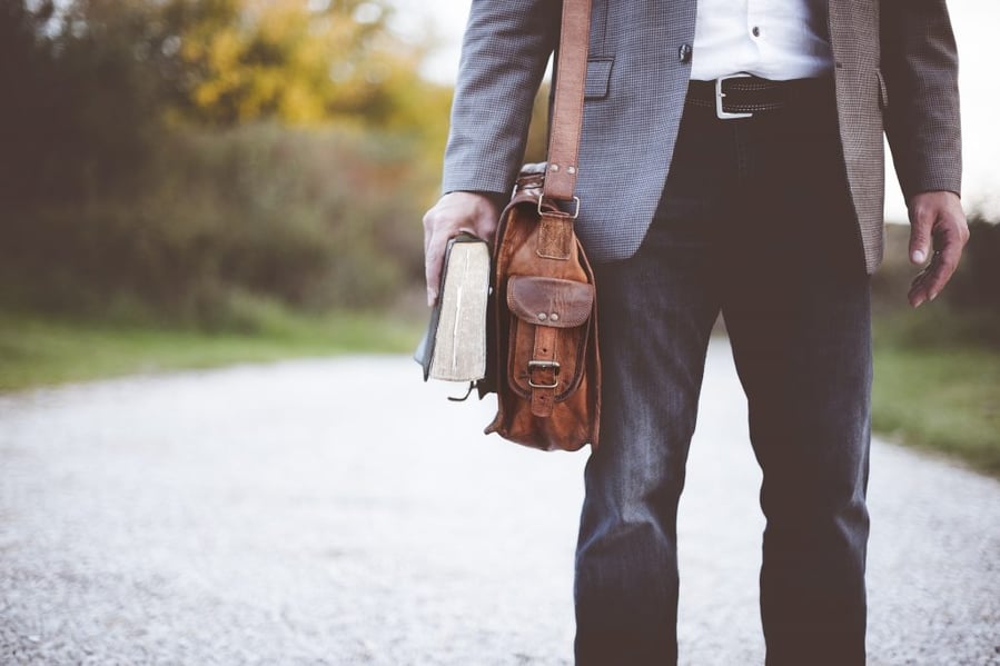 Man standing on a gravel path holding a worn book in one hand and a brown leather shoulder bag in the other, dressed in jeans and a blazer.