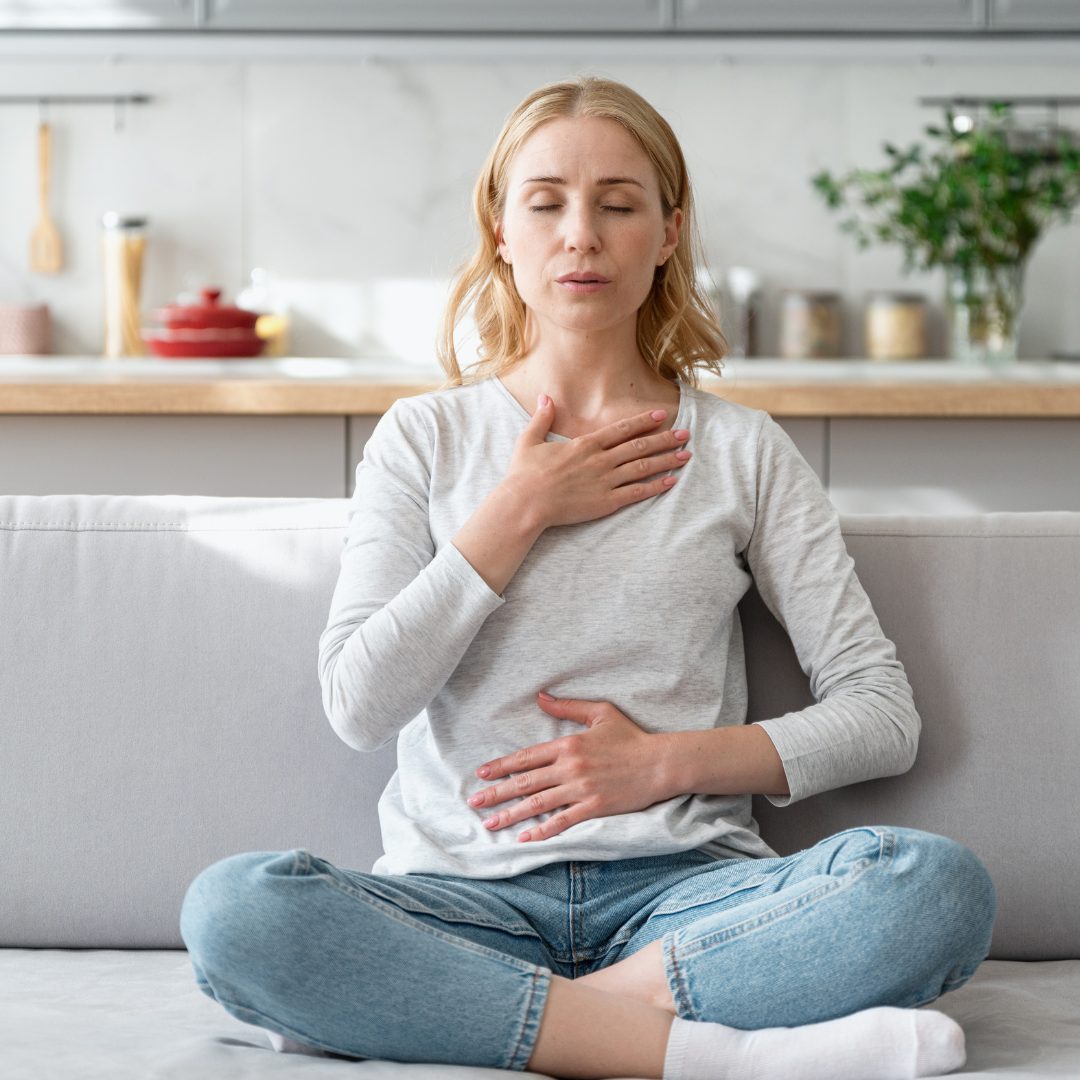 A woman sitting cross-legged on a light-colored sofa, with her eyes closed and a distressed expression. She has one hand on her chest and the other on her stomach, indicating she may be experiencing discomfort or pain. The background shows a modern kitchen with various items on the countertop.