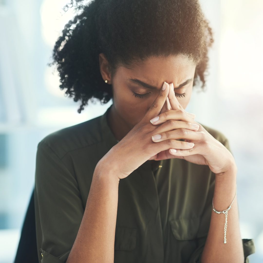 A woman with curly hair is sitting with her eyes closed, her hands pressed together in front of her face, and her fingers resting on her forehead. She appears to be deep in thought or experiencing stress or worry. The background is softly lit and out of focus.