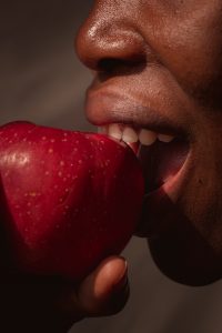 A close-up of a person about to bite into a red apple, holding it with their hand. The person's lips are slightly parted, and their teeth are visible as they prepare to take a bite. The skin tone is dark, and the focus is on the apple and the person's mouth.