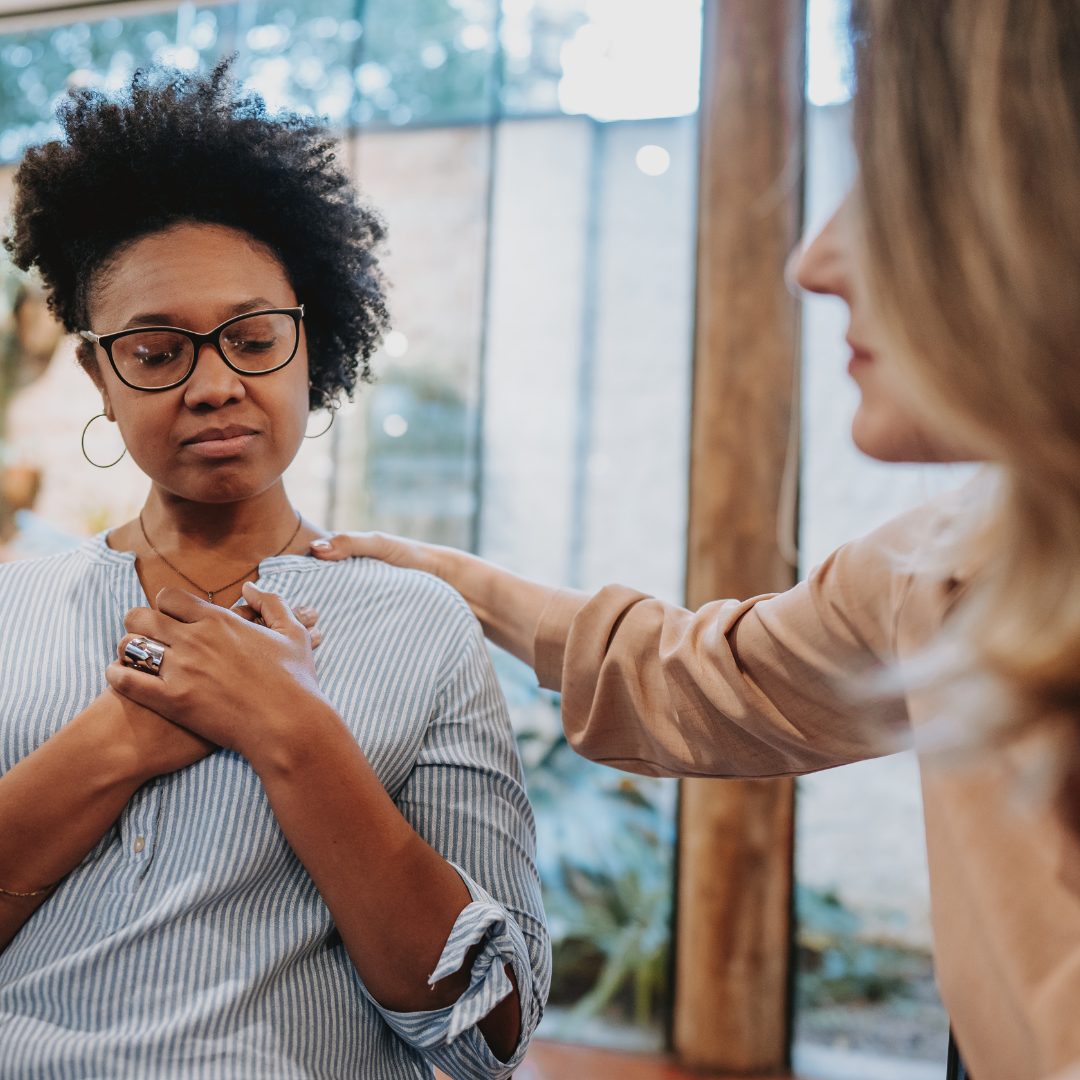 A woman with curly hair and glasses, wearing a striped shirt, has her hands over her chest and looks down with a serious or contemplative expression. Another woman, partially visible on the right, gently places her hand on the first woman's shoulder, offering comfort or support. The background shows large windows with natural light coming in.