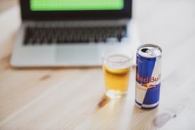 A can of Red Bull energy drink and a glass of beer are placed on a light wooden table in front of a laptop computer with a green screen. The setting suggests a workspace or casual environment, with the focus on the beverages and the open laptop in the background.
