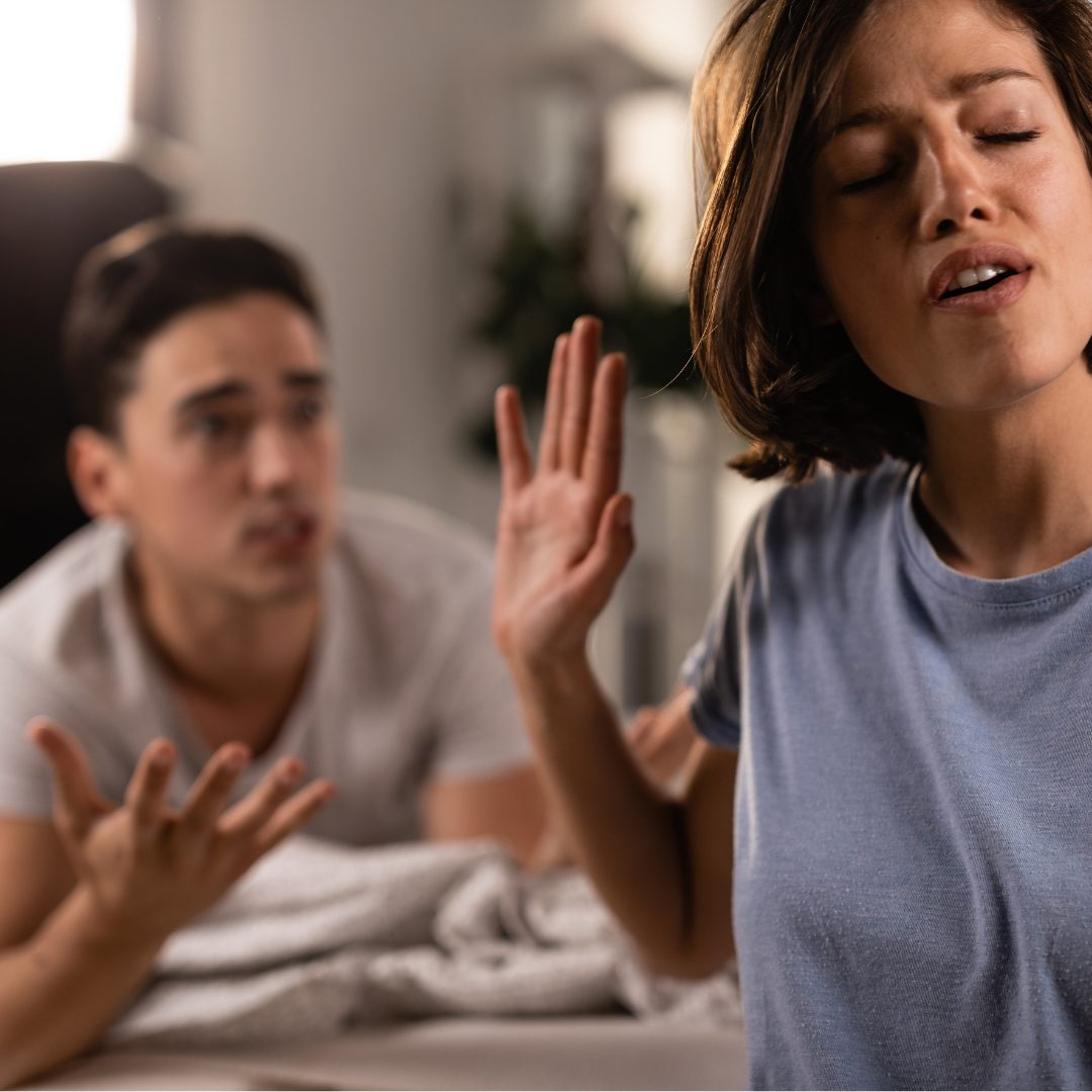 A woman with short brown hair appears to be upset or angry, with her eyes closed and mouth slightly open, raising her hand in a dismissive or defensive gesture. In the background, a man with dark hair and a white shirt looks confused or frustrated, with his hands raised in a questioning or pleading manner. They are indoors, with a blurred background including a bed and a plant.