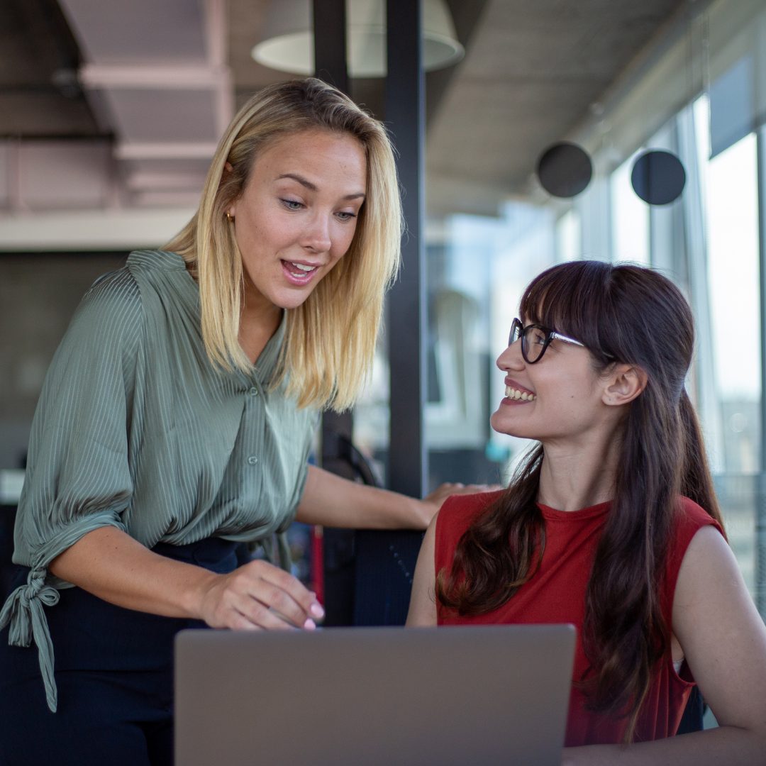 Two women are engaged in a conversation in an office setting. The woman on the left has blonde hair, is wearing a green blouse, and is leaning over a laptop, speaking animatedly. The woman on the right has long dark hair, glasses, and is wearing a red sleeveless top, smiling and looking up at the other woman. They appear to be having a friendly and lively discussion.