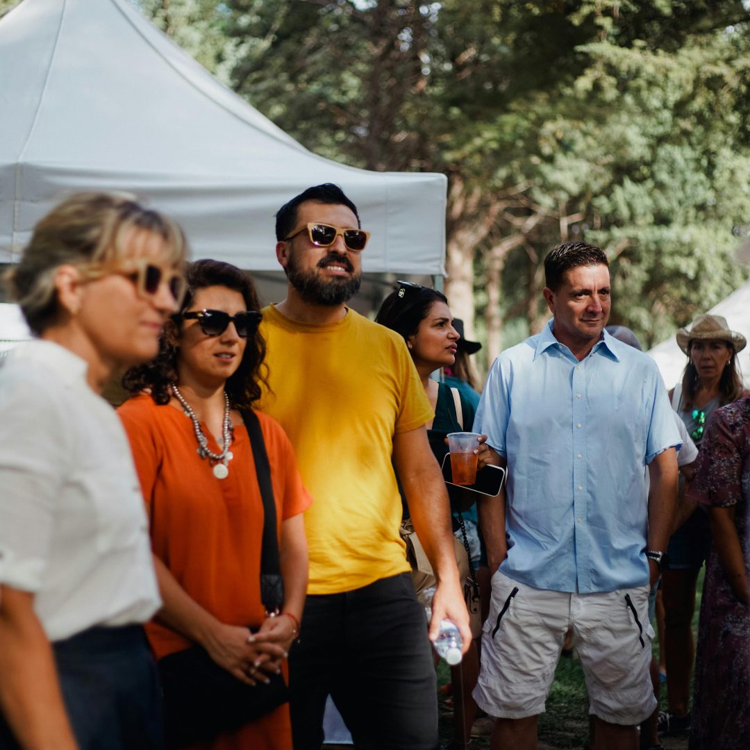 A group of people standing outdoors at an event with white tents and trees in the background. The group includes a woman with blonde hair and sunglasses, a woman with curly dark hair wearing a red-orange top and a pearl necklace, a man with a beard and sunglasses wearing a yellow shirt, and a man in a light blue shirt holding a drink. They all appear to be listening or watching something attentively.