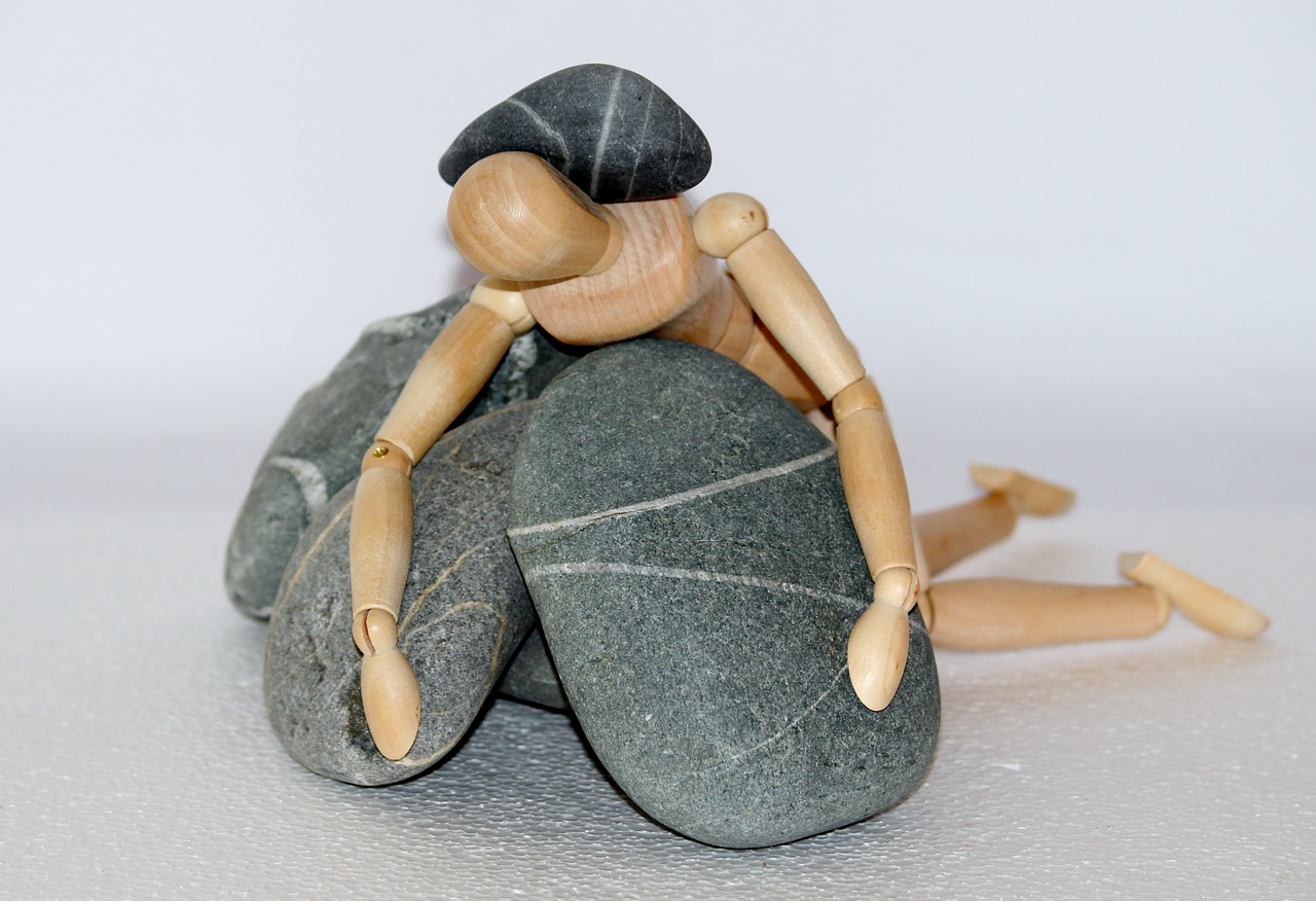Wooden mannequin figure slumped over large gray stones, symbolizing heaviness, exhaustion, or feeling weighed down by emotional or mental burdens.