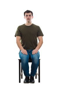 Man sitting upright on a chair with hands resting on his thighs, maintaining a neutral posture as if preparing for meditation or a grounding exercise.